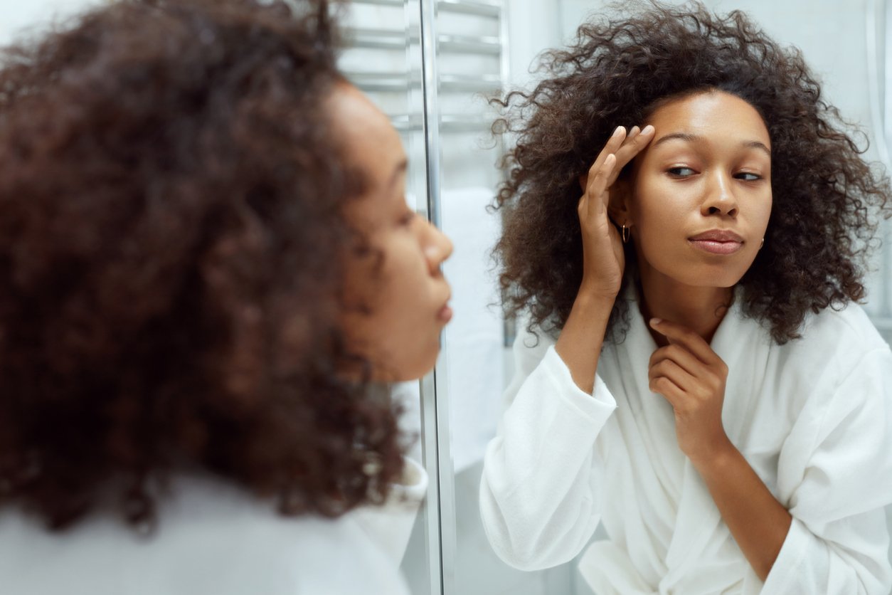 Skin care. Woman touching face skin and looking at mirror at bathroom. Portrait of beautiful african girl with afro hair after shower going through her morning beauty routine