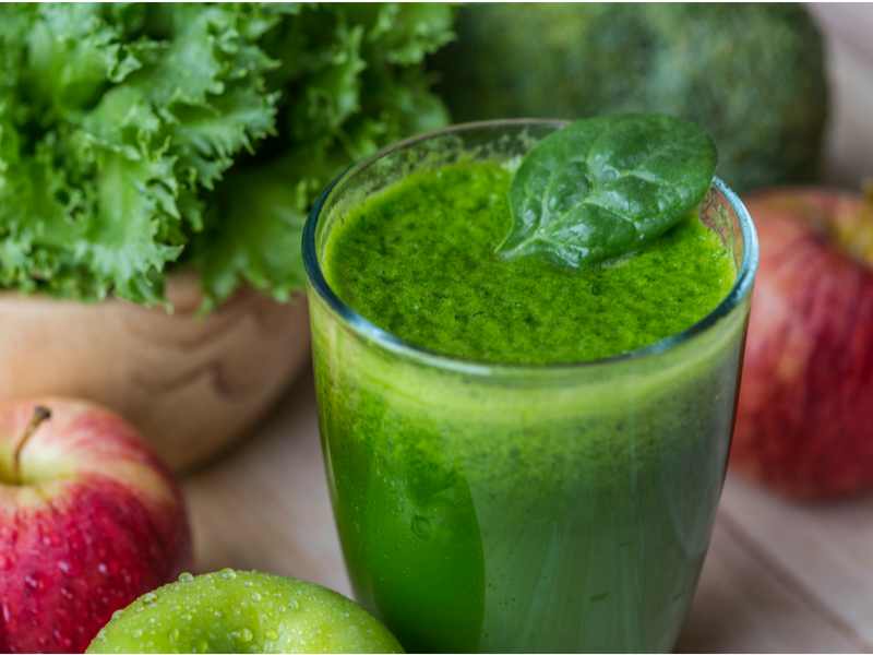 A green drink next to a green apple, a red apple, and some leafy green herb that may or may not be cilantro.