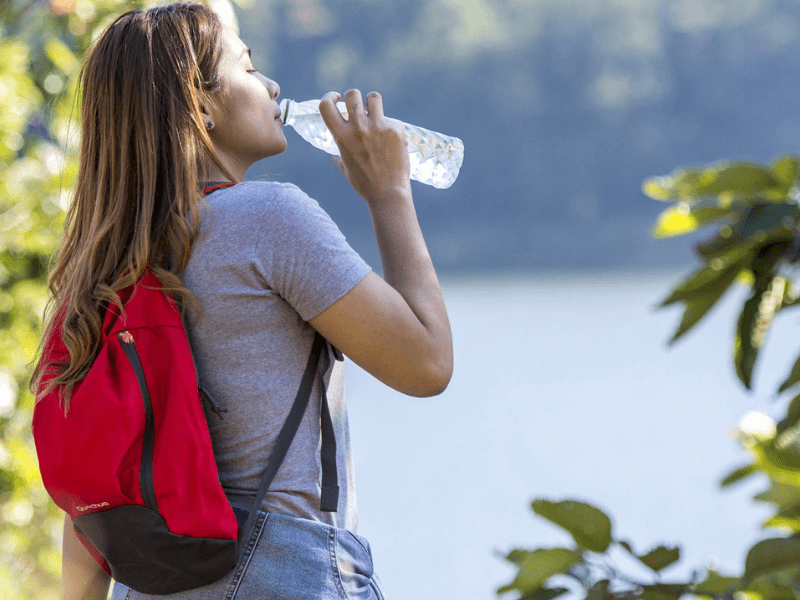 A woman standing by a lake drinking water while doing a whole body detox