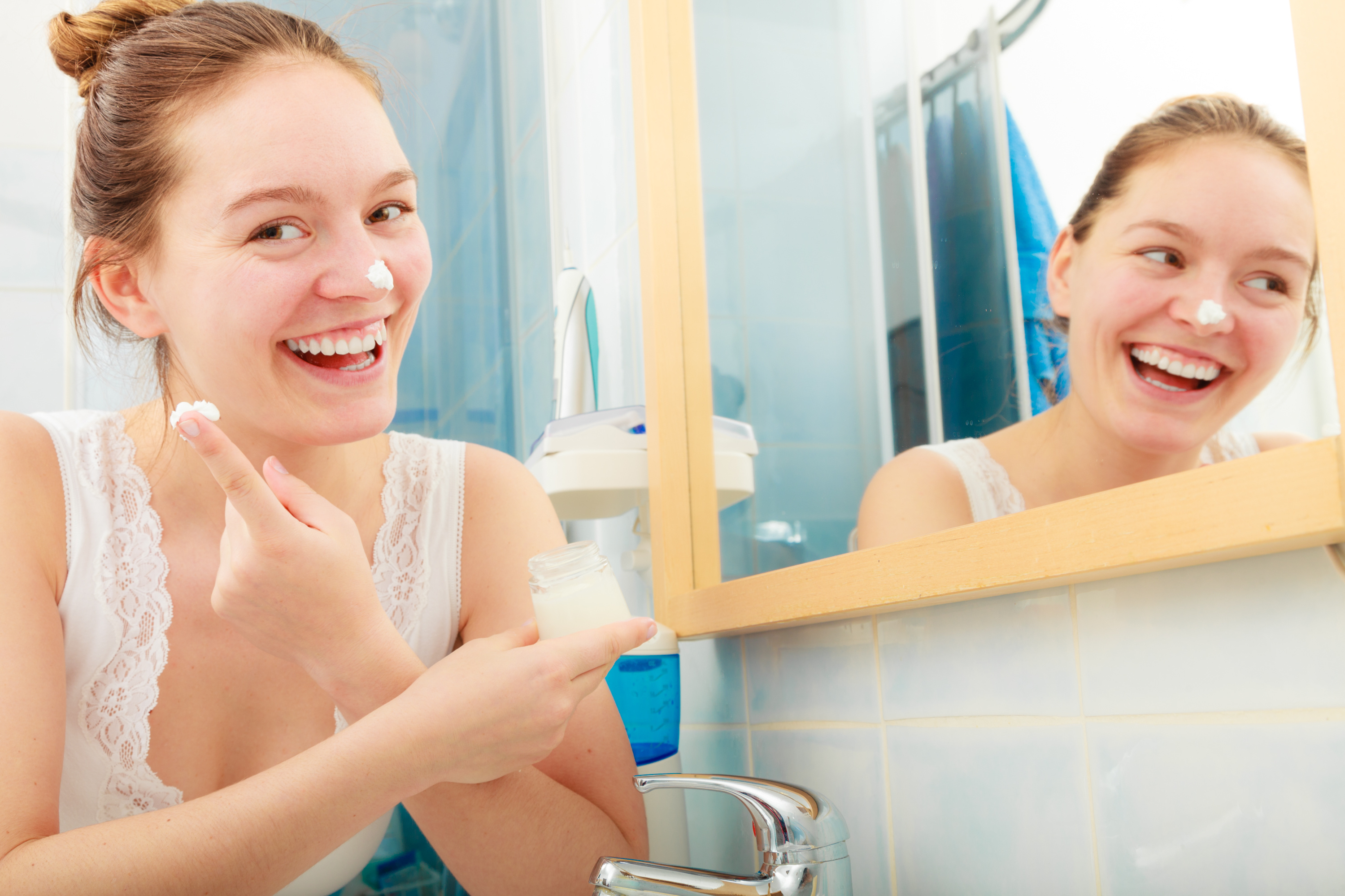 Happy young woman applying cleansing moisturizing skin cream on face. Girl taking care of dry complexion layering moisturizer. Skincare.