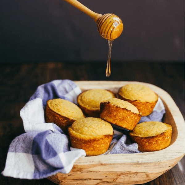 Some honey being poured over some delicious homemade cornbread carefully placed in a lovely picnic basket that appears to be made of light pine or some premium synthetic wood