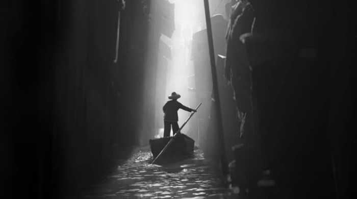 The Streets of 1950s Hong Kong Fan Ho: A man steering a small boat through a dark, narrow channel with high walls.