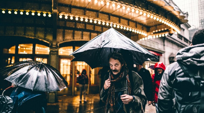 The Streets of New York City by Shaquel Munroe: Pedestrians walking in the rain with umbrellas under bright lights.