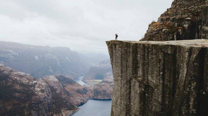 Alex Strohl Fjords of Norway: A small figure standing on the edge of a sheer cliff overlooking the fjords and lakes.