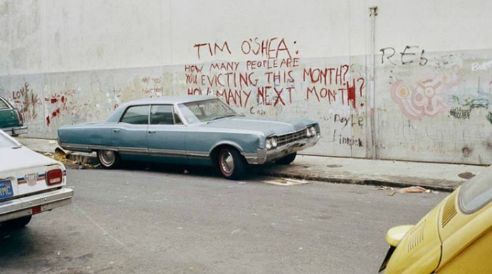 1970s San Francisco by Janet Delaney: An classic, blue car parked against a concrete wall which has graffiti reading "Tim O'Shea: How many people are you evicting this month? How many next month?"