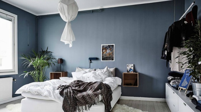A bedroom incorporating the dark wall interior trend with dark blue walls, contrasted with a white ceiling, white bed and light floorboards.