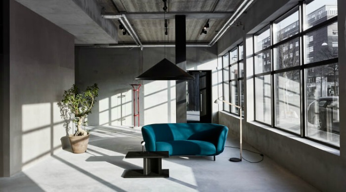 A concrete room in an industrial building set up by Massproductions with a large potted plant, turquoise sofa, standing lamp, black table and large black hanging lamp at the Stockholm Furniture and Light Fair 2016.