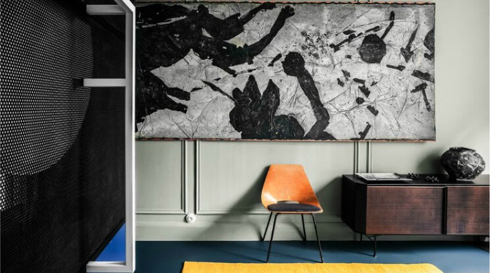 A large black and white modern art painting on a wall with traditional wall moldings, a bright yellow carpet and an orange chair on a dark blue floor in a 1930s apartment.