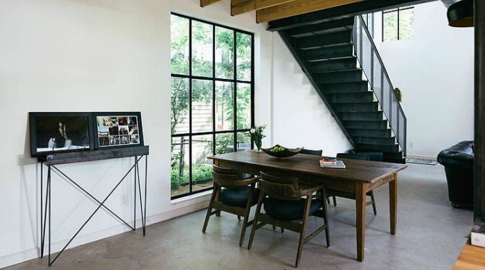 A monochrome living area with white walls, a black staircase and chairs, grey floowing and a dark brown wooden table.