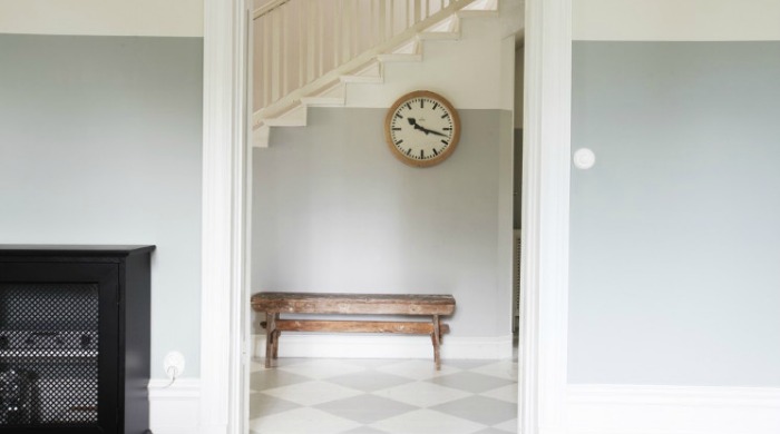 The hallway in a Swedish country house with pastel coloured walls, grey and white tiled floors, a rustic wooden bench and a clock.