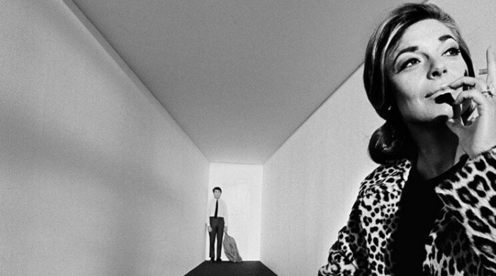 The Art of Behind the Scenes: Anne Bancroft in the foreground of a corridor smoking a cigarette with Dustin Hoffman in the background on the set of 'The Graduate'.