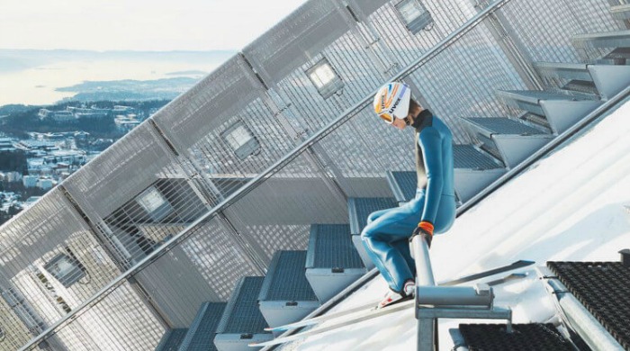 A ski jumper in a blue one-piece with helmet and skis on at the top of the ski slope preparing to take off in the Skihopp series by David Ryle.