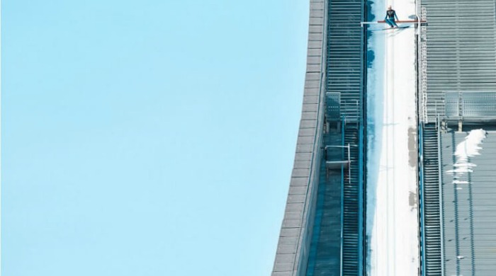 A ski jumper at the top of the slope dwarfed by the size and height of it against a blue sky in the Skihopp series by David Ryle.