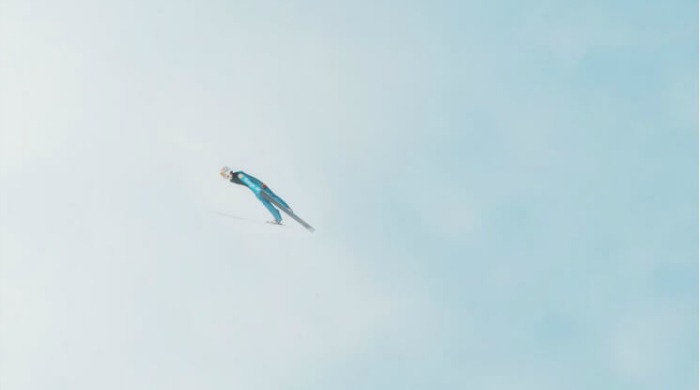 An action shot of a ski jumper in the air, leaning forwards against a blue sky and white cloud in the Skihopp series by David Ryle.