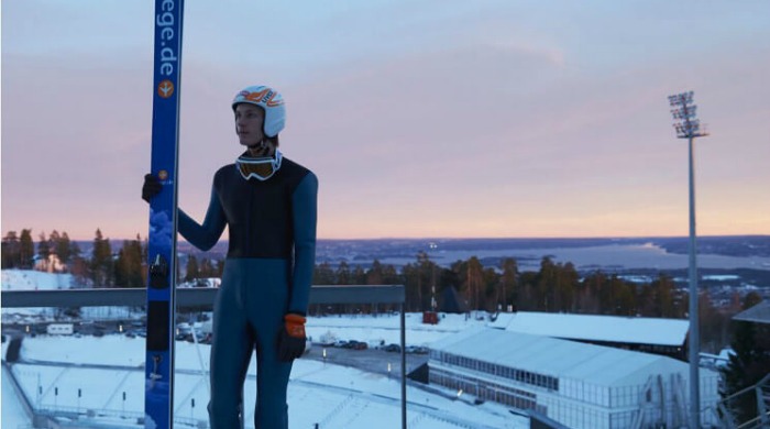 A ski jumper in a blue one-piece with a helmet on holding the skis in front of a sunset in the background in the Skihopp series by David Ryle.