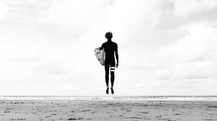A black and white shot of a surfer holding a surfboard under one arm and jumping into the air in the Ocean Culture series by Luke Paige.