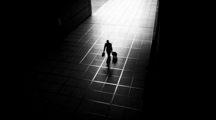 A man walking across tiled flooring with a drag along suitcase lit by a spotlight and surrounded by darkness by Junichi Hakoyama.