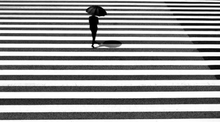 A woman dressed in black holding an umbrella walking across an area of narrow black and white stripes by Junichi Hakoyama.