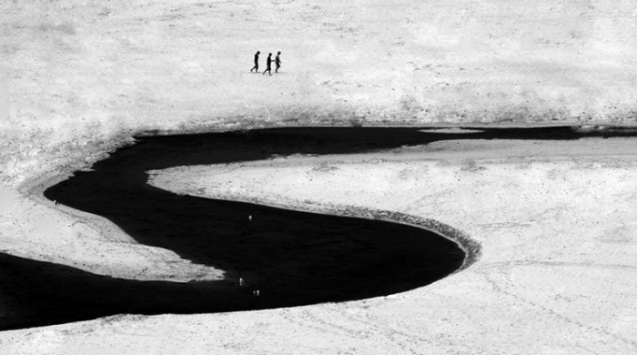 Three small figures walking by a large, black river by Kjetil Karlsen.