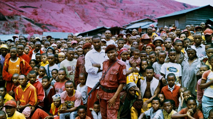 A crowd of civilians and a soldier in front of a pink hillside from 'Infra' by Richard Mosse.