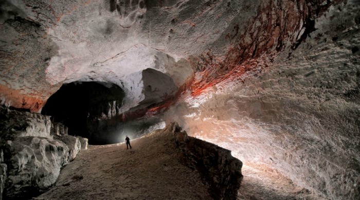 A caver in a large cave with red streaks along the walls by Robbie Shone.