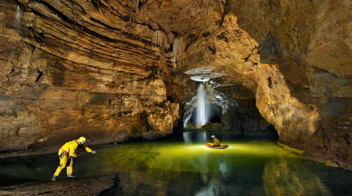 Cavers crossing a yellow-lit underground lake by Robbie Shone.