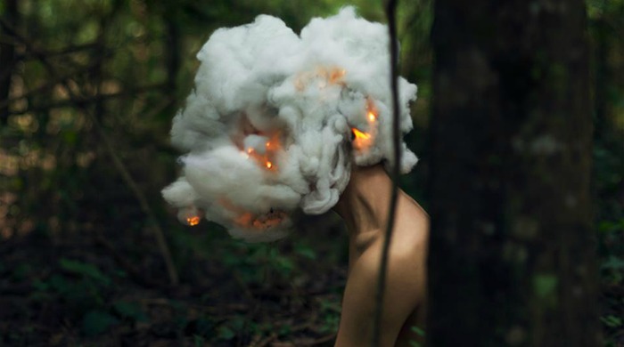 A woman in a forest with flames and a cloud of smoke covering her head by Mikael Aldo.