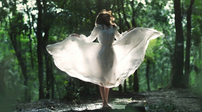 A woman in a forest wearing a floating, white dress by Mikael Aldo.